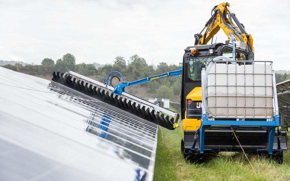 SunBrush® Rapid montado en una miniexcavadora con tanque de agua, limpiando módulos de una planta fotovoltaica en suelo